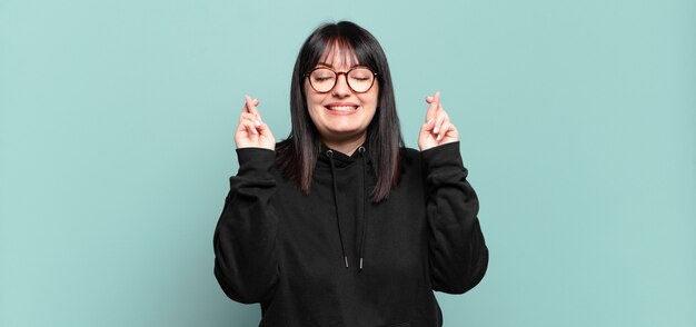 Mulher bonita plus size sorrindo e cruzando os dedos ansiosamente, preocupada e desejando ou esperando boa sorte