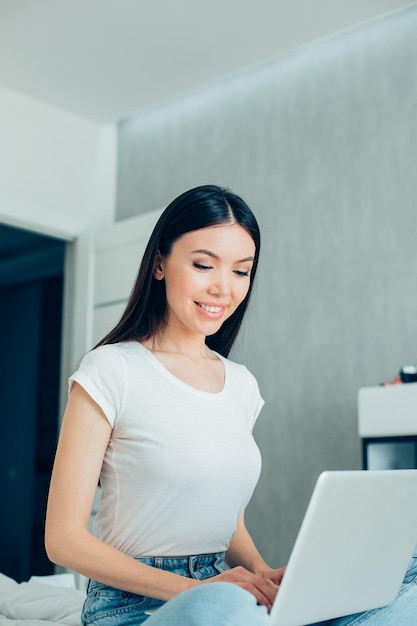 Mulher bonita positiva em roupas casuais sentada com um laptop moderno no colo e olhando para a tela com um sorriso