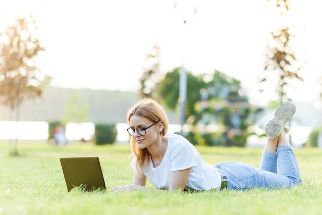 Mulher bonita que trabalha com o laptop no parque na grama. O conceito de trabalho remoto