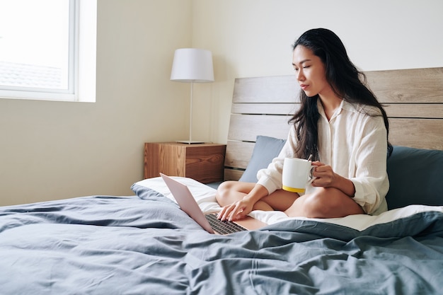 Mulher bonita que trabalha em casa devido a uma pandemia. Ela está sentada na cama, bebendo café e checando e-mails