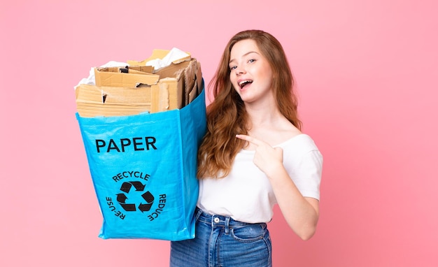 Mulher bonita ruiva, animada e surpresa, apontando para o lado e segurando uma sacola de papel reciclado