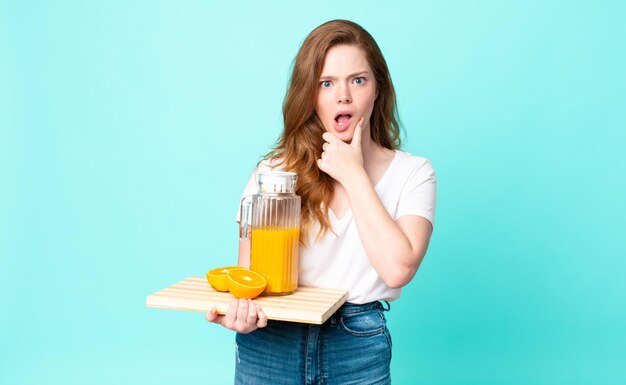 Mulher bonita ruiva com boca e olhos bem abertos e mão no queixo segurando um suco de laranja