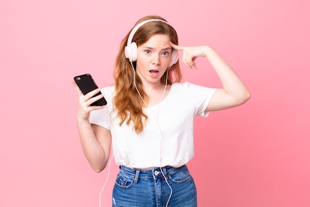 Mulher bonita ruiva parecendo surpresa, percebendo um novo pensamento, ideia ou conceito com fones de ouvido e smartphone