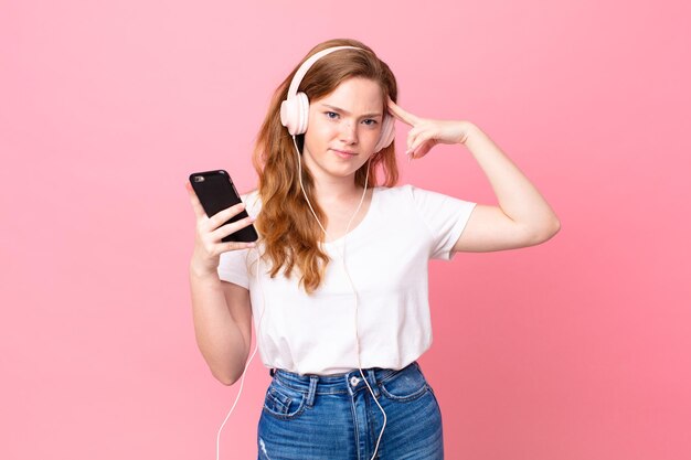 Mulher bonita ruiva se sentindo confusa e intrigada, mostrando que você é louco com fones de ouvido e smartphone