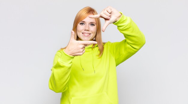 Mulher bonita ruiva se sentindo feliz, amigável e positiva, sorrindo e fazendo um retrato ou moldura com as mãos