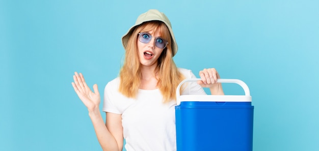 Mulher bonita ruiva se sentindo feliz, surpresa ao perceber uma solução ou ideia e segurando uma geladeira portátil. conceito de verão