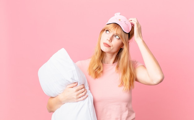 Mulher bonita ruiva sentindo-se perplexa e confusa, coçando a cabeça. vestindo pijama e segurando um travesseiro