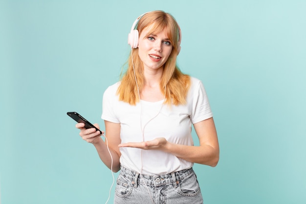 Mulher bonita ruiva sorrindo alegremente, se sentindo feliz e mostrando um conceito e ouvindo música com fones de ouvido