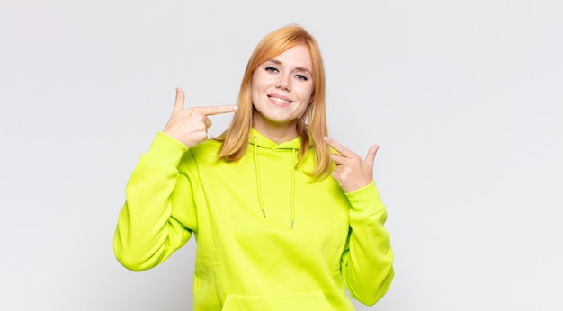 Mulher bonita ruiva sorrindo com confiança apontando para o próprio sorriso largo, atitude positiva, relaxada e satisfeita