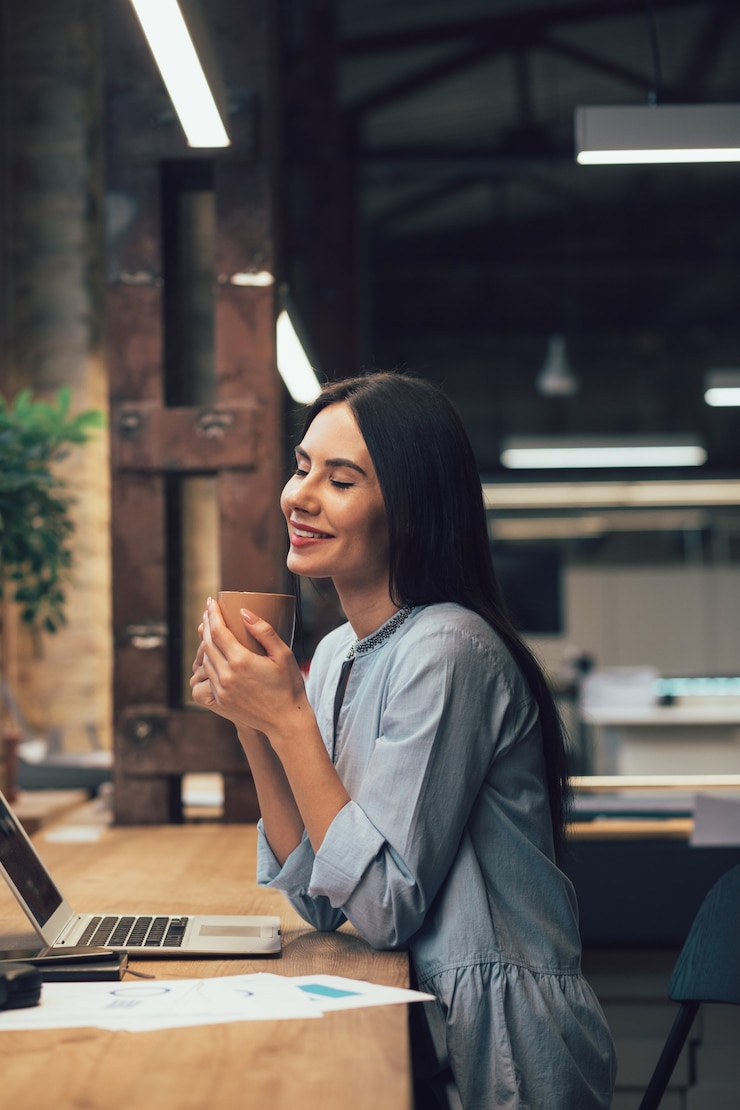 Foto mulher bonita satisfeita segurando uma caneca e sorrindo com os olhos fechados enquanto está sentada à mesa do escritório