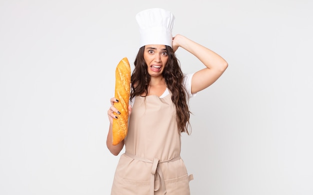 Mulher bonita se sentindo estressada, ansiosa ou com medo, com as mãos na cabeça usando um avental e segurando uma baguete de pão