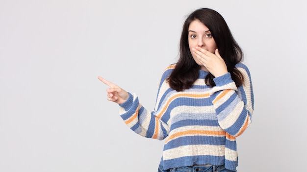 Mulher bonita se sentindo feliz, chocada e surpresa, cobrindo a boca com a mão e apontando para o espaço lateral da cópia