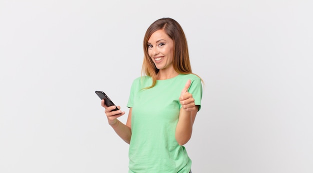 Mulher bonita se sentindo orgulhosa, sorrindo positivamente com o polegar para cima e usando um smartphone