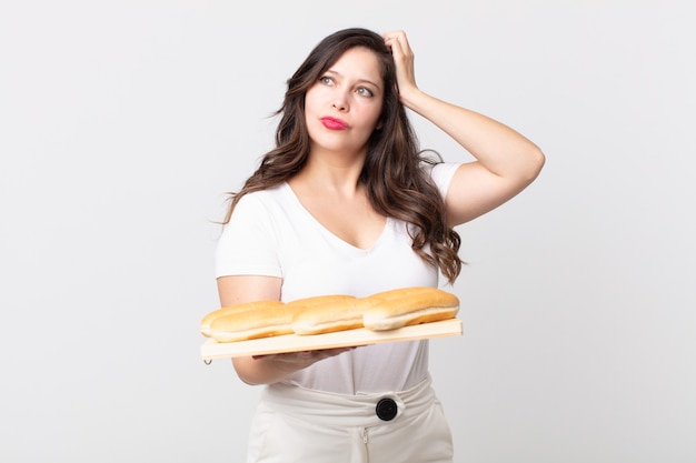 Mulher bonita se sentindo perplexa e confusa, coçando a cabeça e segurando uma bandeja de pão