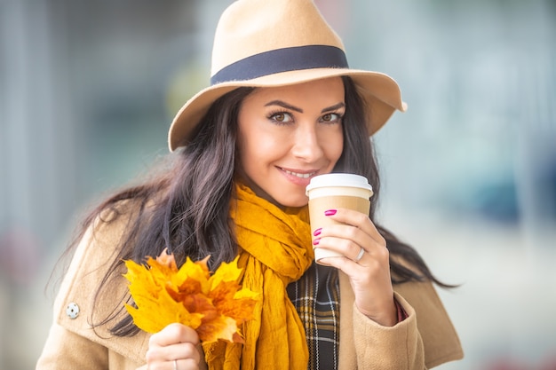 Mulher bonita segura uma xícara de café para viagem e um monte de folhas coloridas colhidas.