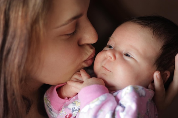 Mulher bonita segurando o bebê recém-nascido em seus braços beijando sua pequena bochecha suavemente enquanto posa em casa, a mãe desfrutando de doces momentos felizes com o bebê