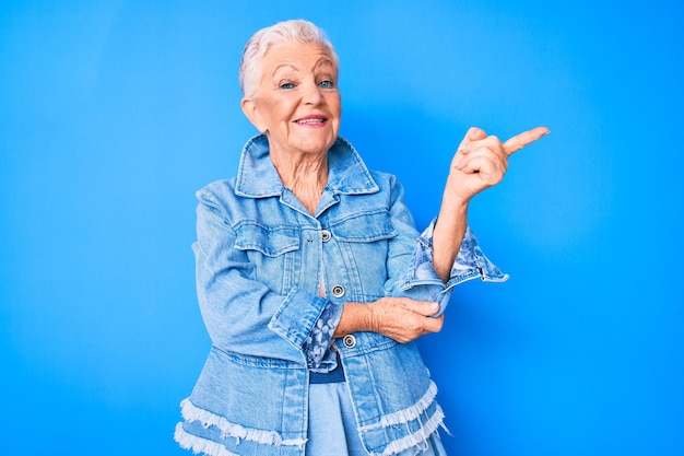 Foto mulher bonita sênior com olhos azuis e cabelos grisalhos vestindo jeans estilo casual sorrindo feliz apontando com a mão e o dedo para o lado