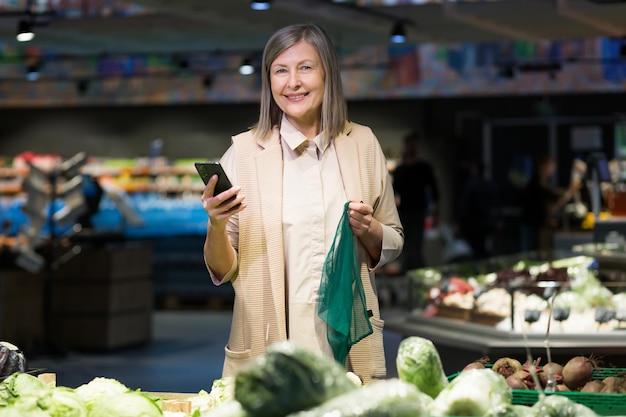 Mulher bonita sênior comprando legumes na loja Supermercado Ele fica segurando o telefone nas mãos segura a rede de alimentos escolhe Ele olha para a câmera sorri