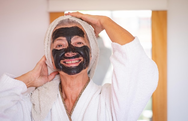 Mulher bonita sênior despreocupada com uma máscara de carvão facial de desintoxicação caseira sorrindo olhando para a câmera cuidar do conceito de pele