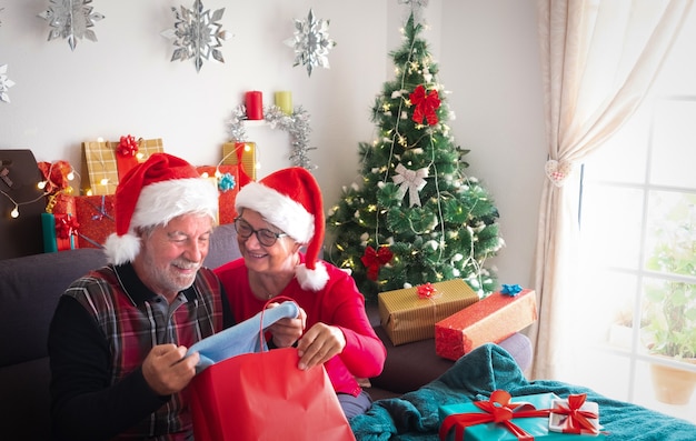 Mulher bonita sênior olha para o marido, dando-lhe um belo suéter azul como presente de Natal. Muitos pacotes de presentes perto deles para a família e amigos