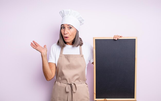 Mulher bonita sênior olhando surpreso e chocado, com o queixo caído segurando um objeto. conceito de chef e quadro-negro