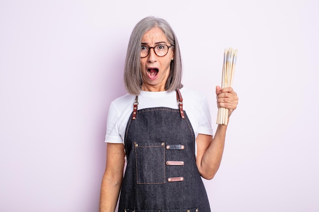 Mulher bonita sênior parecendo muito chocada ou surpresa. artista com pincéis