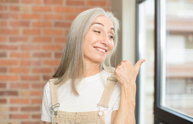 Mulher bonita sênior sorrindo alegre e casualmente apontando para copiar o espaço ao lado, sentindo-se feliz e satisfeita