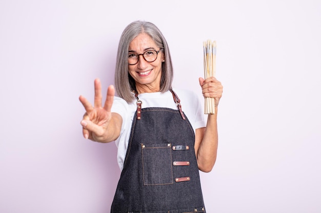 Mulher bonita sênior sorrindo e parecendo amigável, mostrando o número três. artista com pincéis