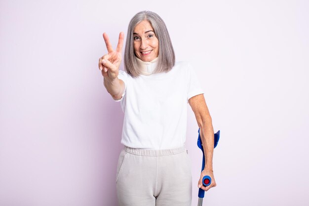 Mulher bonita sênior sorrindo e parecendo feliz gesticulando vitória ou muleta de paz e conceito de acidente de colar