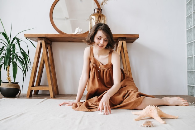 Mulher bonita sensual curiosa sentada no chão com um vestido marrom, tocando uma concha. Bela morena com cabelo curto dentro de casa.