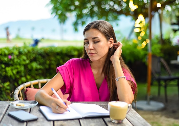 Mulher bonita sentada em uma cadeira no jardim de verão e escrevendo em um caderno