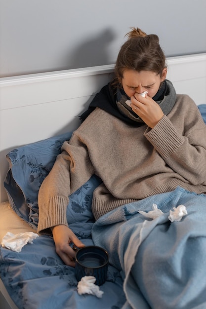 Mulher bonita sentada na cama, assoa o nariz em um guardanapo de papel e segura a xícara com chá quente. Menina com coriza, rinite ou sinusite. Doença sazonal. Tratamento em casa. Conceito de saúde