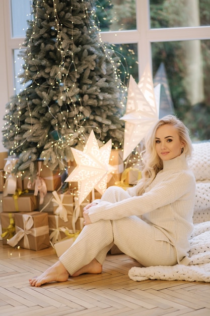 Mulher bonita sentada no parapeito da janela perto da árvore de Natal. Mulher de cabelo loiro com fantasia branca descanse