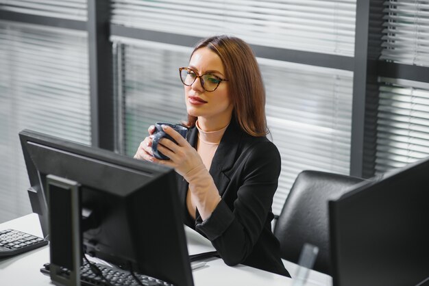 Mulher bonita, simpática, fofa e perfeita, sentada em sua mesa na cadeira de couro na estação de trabalho, usando óculos, trajes formais, com laptop e notebook na mesa