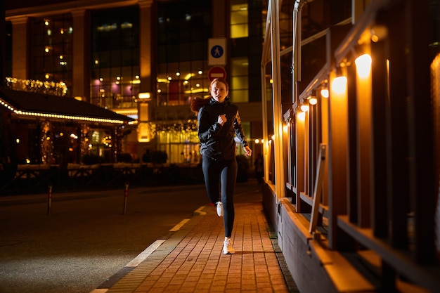 Mulher bonita slim fitness em roupa esportiva preta está correndo na cidade à noite rua feminino está fazendo ...