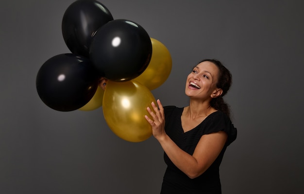 Foto mulher bonita sorridente alegre segurando balões de ar inflados brilhantes ouro preto contra o fundo da parede cinza com espaço de cópia. balões dourados - conceito de cartão de crédito ouro. balões pretos - black friday