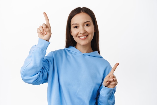 Mulher bonita sorridente apontando para o lado, mostrando duas opções, variantes para clientes, em pé em roupas casuais contra uma parede branca.