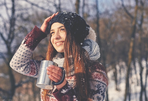 Mulher bonita sorridente com copo de bebida quente em pé perto da floresta de inverno