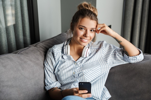 Mulher bonita sorridente com fones de ouvido usando o celular enquanto está sentada no sofá na sala de estar