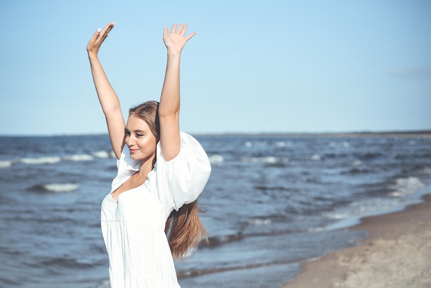 Mulher bonita sorridente feliz na praia do oceano em pé em um vestido branco de verão, levantando as mãos