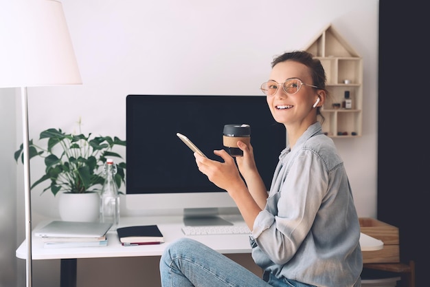 Mulher bonita sorridente moderna trabalhando e bebendo café da caneca reutilizável no escritório em casa Local de trabalho minimalista de estilo de vida lento de mulher criativa em óculos e fones de ouvido usando tecnologia de computador