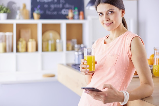 Mulher bonita sorridente olhando para o celular e segurando o copo de suco de laranja enquanto toma café da manhã em uma cozinha