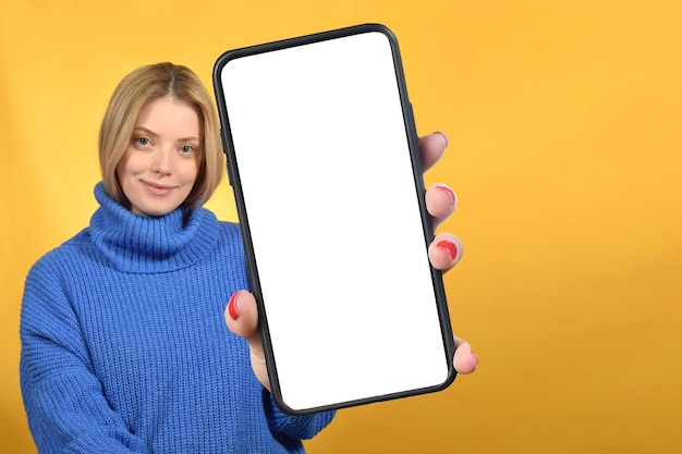 Mulher bonita sorridente segurando smartphone com tela de dispositivo branco em branco na mão perto da câmera