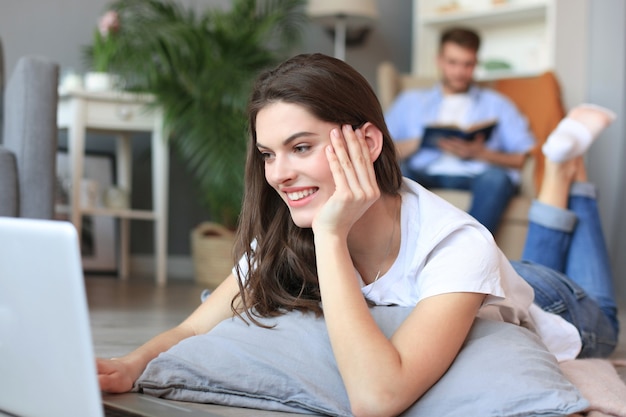 Mulher bonita sorridente usando laptop com homem borrado no fundo em casa.