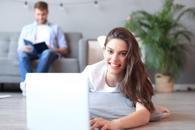 Mulher bonita sorridente usando laptop com homem borrado no fundo em casa.