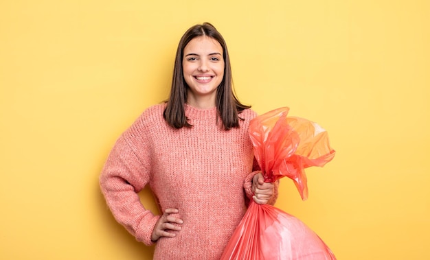 Mulher bonita sorrindo alegremente com uma mão no quadril e confiante. conceito de saco de lixo