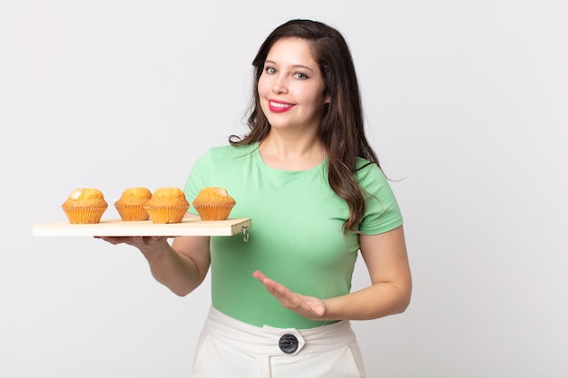 Mulher bonita sorrindo alegremente, se sentindo feliz, mostrando um conceito e segurando uma bandeja de muffins