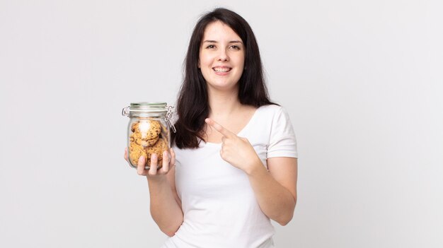 Mulher bonita sorrindo alegremente, sentindo-se feliz e apontando para o lado segurando uma garrafa de vidro de biscoitos