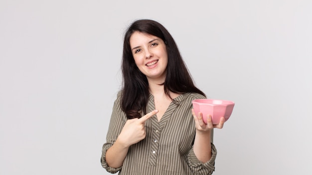 Mulher bonita sorrindo alegremente, sentindo-se feliz e apontando para o lado segurando uma tigela de panela vazia. agente assistente com fone de ouvido