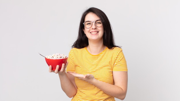 Mulher bonita sorrindo alegremente, sentindo-se feliz, mostrando um conceito e segurando uma tigela de café da manhã